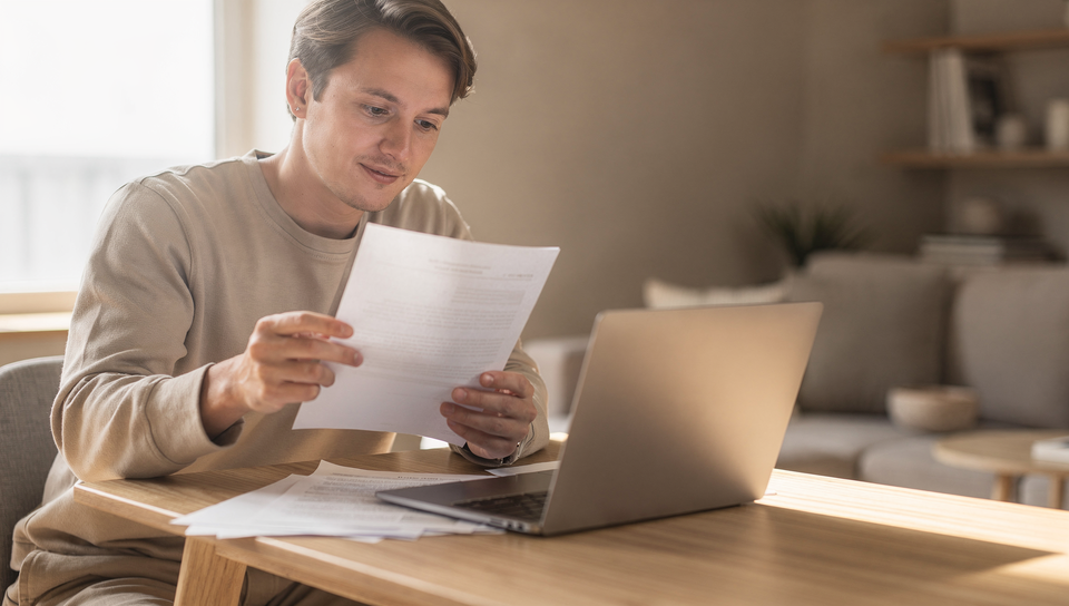 Person prüft am Schreibtisch ein Dokument neben dem Laptop – Grundbuchauszug kostenlos oder kostenpflichtig?